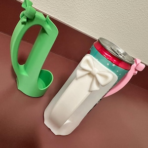 May include: A green 3D-printed can holder and a white bow-shaped can sleeve. The can sleeve is wrapped around a red, teal, and silver can. The can holder has a handle and a small cup to hold the can.