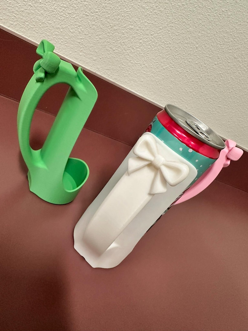 May include: A green 3D-printed can holder and a white bow-shaped can sleeve. The can sleeve is wrapped around a red, teal, and silver can. The can holder has a handle and a small cup to hold the can.