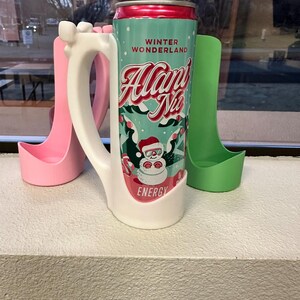May include: A teal and red can of "Alan's Nio Energy" with a Santa Claus graphic, placed in a white mug-shaped holder. Two other holders, one pink and one green, are also visible. The can reads "Winter Wonderland".