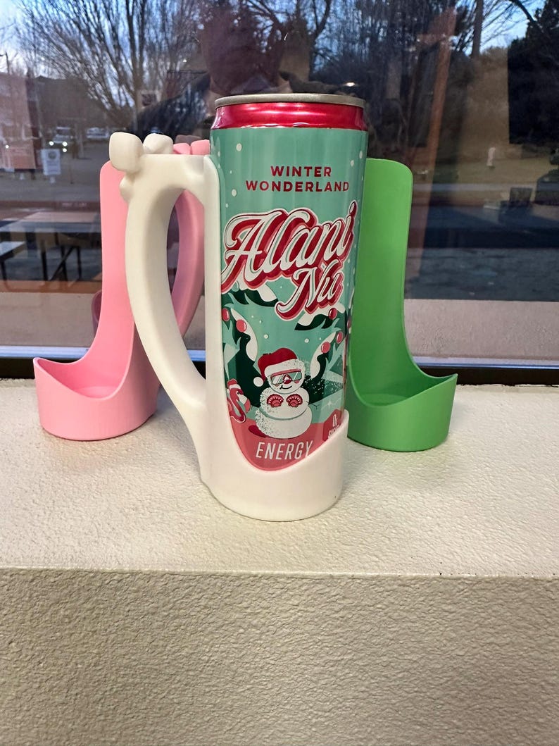 May include: A teal and red can of "Alan's Nio Energy" with a Santa Claus graphic, placed in a white mug-shaped holder. Two other holders, one pink and one green, are also visible. The can reads "Winter Wonderland".