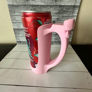 May include: A pink can holder with a handle, designed to fit a standard beverage can. The can is red with a "Berry Lush" label and cherry graphics. The holder is made of plastic and has a smooth finish.