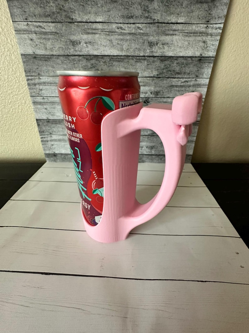 May include: A pink can holder with a handle, designed to fit a standard beverage can. The can is red with a "Berry Lush" label and cherry graphics. The holder is made of plastic and has a smooth finish.