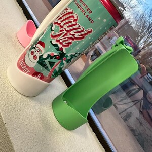 May include: A can of "Winter Wonderland" energy drink in a white and green holder, with a pink and green holder nearby. The can has a red top and features a snowman graphic. The text "Winter Wonderland" and "Hans Ne" are visible.