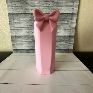 May include: A pink, geometric-shaped vase with a bow on top. The vase has a faceted design and is a soft, pastel pink color. The bow is the same color as the vase. The vase is sitting on a white wooden surface.