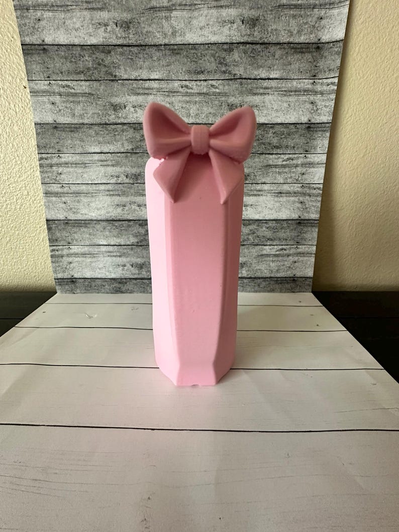 May include: A pink, geometric-shaped vase with a bow on top. The vase has a faceted design and is a soft, pastel pink color. The bow is the same color as the vase. The vase is sitting on a white wooden surface.