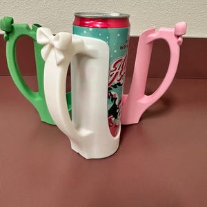 May include: Three colorful can holders in green, white, and pink, each with a handle. The white holder has a bow detail. A can with a red and teal label is placed inside the white holder. The holders are on a maroon surface.