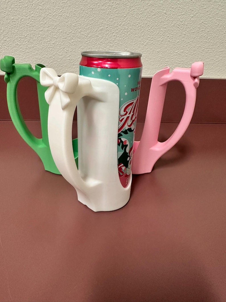 May include: Three colorful can holders in green, white, and pink, each with a handle. The white holder has a bow detail. A can with a red and teal label is placed inside the white holder. The holders are on a maroon surface.