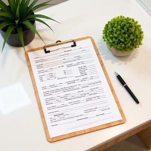 May include: A wooden clipboard with a gold-coloured clip holding a white form with black text. A black pen rests on the white surface. A potted green plant and a small green ball-shaped plant are also on the white surface.