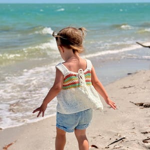 May include: A white crochet top with a rainbow stripe design on the back, paired with denim shorts. The top has a square neckline and a tie closure. The child is standing on a sandy beach with the ocean in the background.