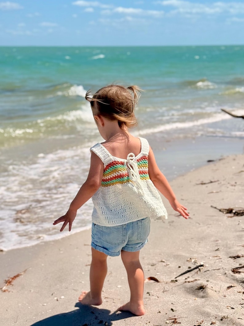 May include: A white crochet top with a rainbow stripe design on the back, paired with denim shorts. The top has a square neckline and a tie closure. The child is standing on a sandy beach with the ocean in the background.