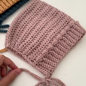 Puede incluir: Primer plano de un gorro de ganchillo rosa claro hecho a mano, con un patr&oacute;n de puntada texturizada. El gorro tiene forma redondeada y lazos bajo la barbilla. Otros gorros en verde azulado y mostaza son visibles.