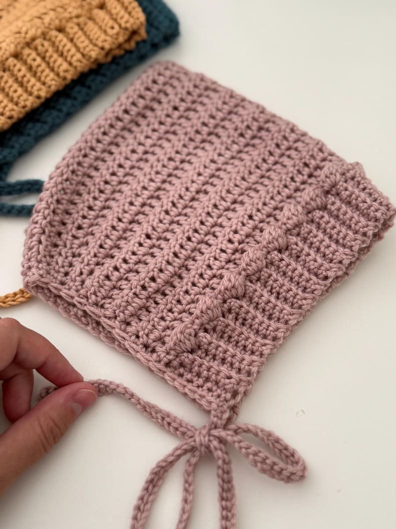 Puede incluir: Primer plano de un gorro de ganchillo rosa claro hecho a mano, con un patr&oacute;n de puntada texturizada. El gorro tiene forma redondeada y lazos bajo la barbilla. Otros gorros en verde azulado y mostaza son visibles.