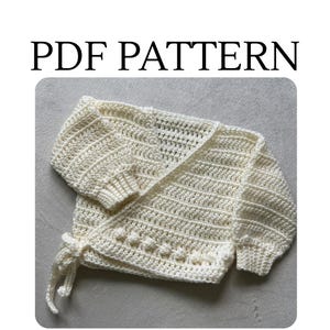 Puede incluir: Un cárdigan cruzado de ganchillo en color crema con mangas largas de puños acanalados y un detalle de puntos de burbuja texturizados en el borde inferior delantero. La prenda incluye un cierre de lazo lateral y dice PDF PATTERN arriba.