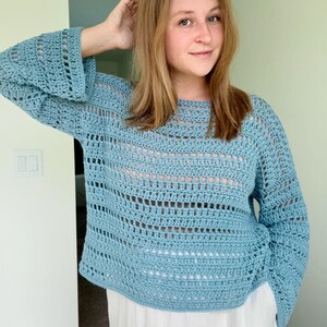 May include: A light blue long-sleeved crochet jumper featuring a loose, open-knit pattern with alternating solid rows and airy, rectangular gaps. The garment has a wide, rounded neckline and slightly flared sleeves, crafted from yarn.