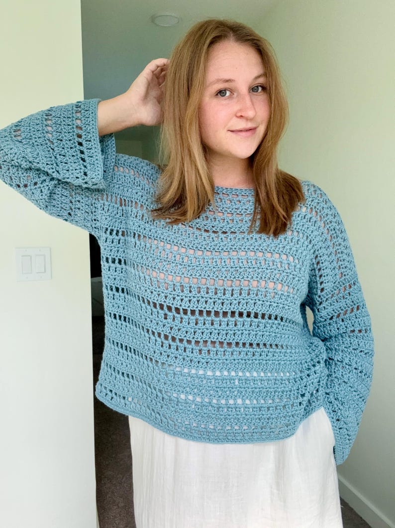 May include: A light blue long-sleeved crochet jumper featuring a loose, open-knit pattern with alternating solid rows and airy, rectangular gaps. The garment has a wide, rounded neckline and slightly flared sleeves, crafted from yarn.
