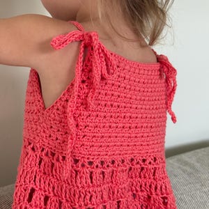 May include: A coral-colored sleeveless top crafted with a crochet technique. The garment features adjustable shoulder straps that tie into bows at the top. The upper section has a dense stitch pattern, while the lower portion transitions into an open, airy, and decorative crochet design with visible gaps.