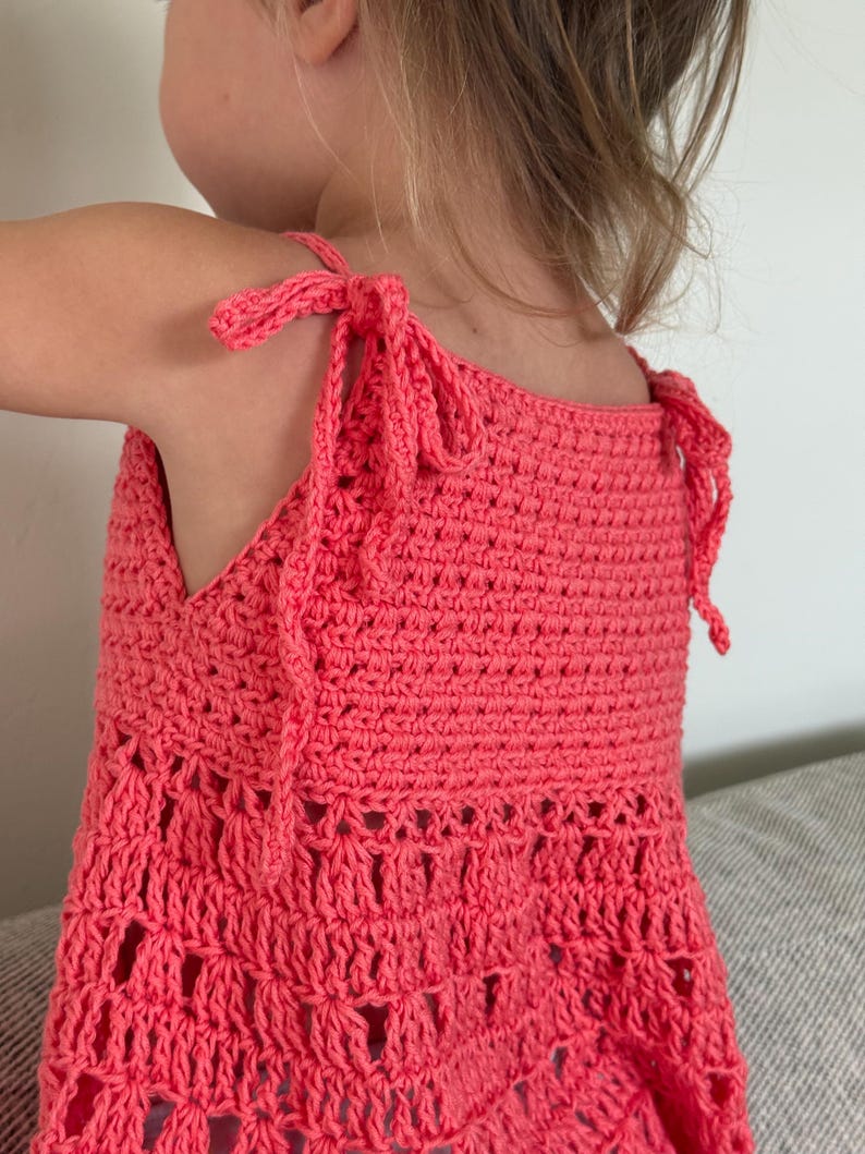 May include: A coral-colored sleeveless top crafted with a crochet technique. The garment features adjustable shoulder straps that tie into bows at the top. The upper section has a dense stitch pattern, while the lower portion transitions into an open, airy, and decorative crochet design with visible gaps.