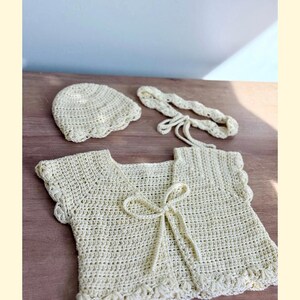 K&ouml;nnte beinhalten: Ein cremefarbenes H&auml;kelset bestehend aus einer kurz&auml;rmeligen Strickjacke mit Bindeband vorne, einer passenden runden M&uuml;tze und einem geflochtenen Stirnband. Jedes Teil weist durchgehend ein zartes Detail mit gewelltem Rand auf.