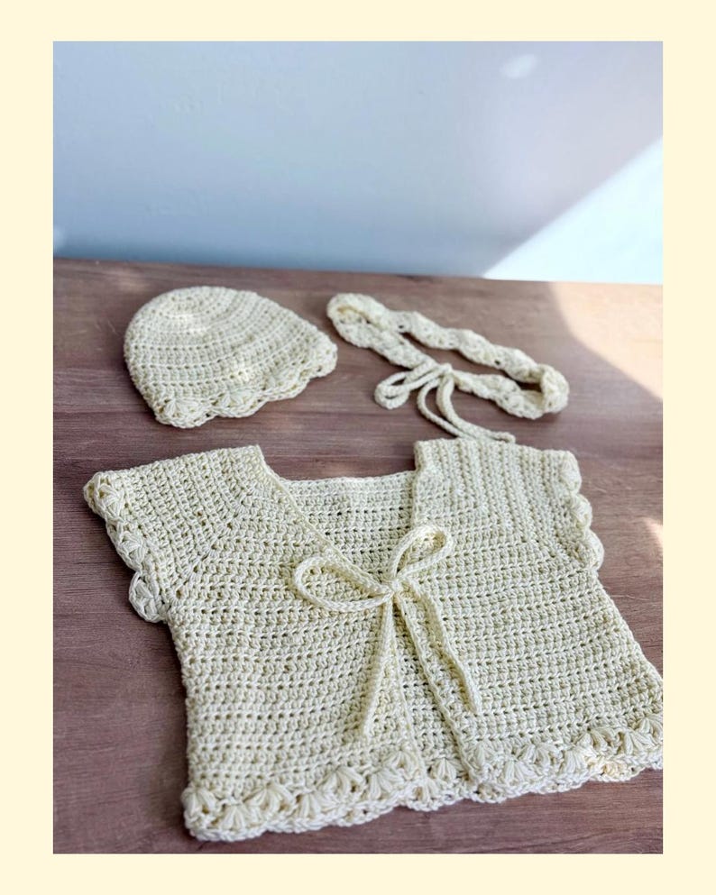 K&ouml;nnte beinhalten: Ein cremefarbenes H&auml;kelset bestehend aus einer kurz&auml;rmeligen Strickjacke mit Bindeband vorne, einer passenden runden M&uuml;tze und einem geflochtenen Stirnband. Jedes Teil weist durchgehend ein zartes Detail mit gewelltem Rand auf.