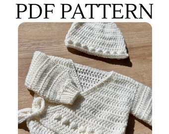 May include: A white crochet PDF PATTERN featuring a long-sleeved wrap cardigan with a textured bobble stitch border and a matching beanie hat. The garment is crafted with a consistent white yarn, showcasing detailed stitch work throughout.