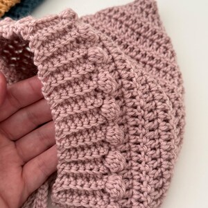 Puede incluir: Un gorro de ganchillo rosa claro hecho a mano con un punto texturizado y detalles de bolitas. El gorro tiene forma redondeada y lazos en la barbilla. El color suave y el dise&ntilde;o intrincado sugieren un art&iacute;culo para beb&eacute;s o ni&ntilde;os.