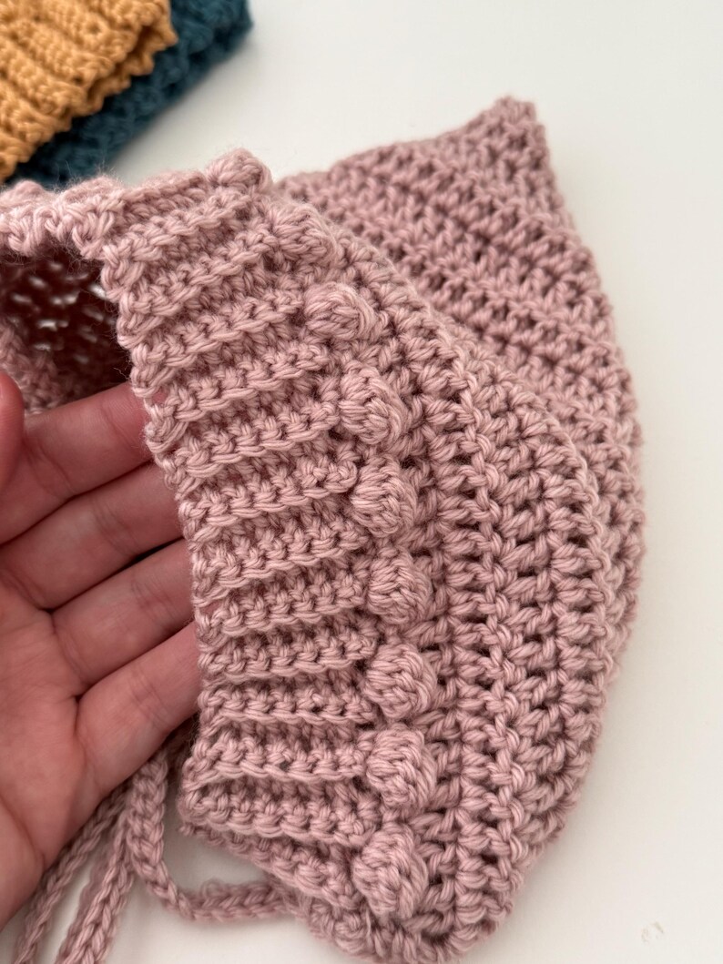Puede incluir: Un gorro de ganchillo rosa claro hecho a mano con un punto texturizado y detalles de bolitas. El gorro tiene forma redondeada y lazos en la barbilla. El color suave y el dise&ntilde;o intrincado sugieren un art&iacute;culo para beb&eacute;s o ni&ntilde;os.