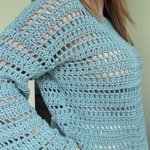 May include: A light blue long-sleeved jumper featuring an openwork crochet pattern with a repeating grid design. The garment has a round neckline and a loose, airy construction with visible gaps between the stitched rows throughout the fabric.