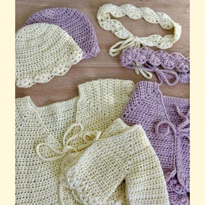 K&ouml;nnte beinhalten: Eine Sammlung handgefertigter H&auml;kelartikel, bestehend aus zwei Strickjacken, zwei M&uuml;tzen und zwei Stirnb&auml;ndern in Creme- und Lavendelt&ouml;nen. Die Strickjacken haben Bindeb&auml;nder, w&auml;hrend die M&uuml;tzen und Stirnb&auml;nder mit Bogenkanten versehen sind.