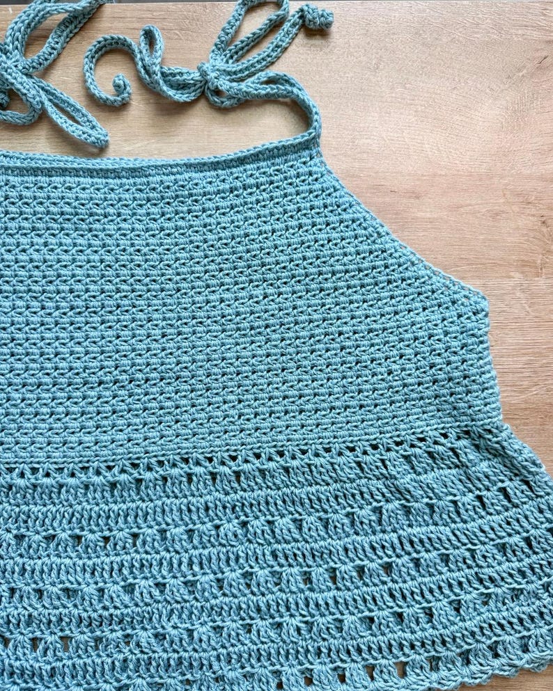 Pu&ograve; includere: Un top all'americana all'uncinetto color azzurro caratterizzato da un motivo a maglia denso e uniforme nella parte superiore e da un motivo decorativo in pizzo traforato sull'orlo svasato, con lacci all'uncinetto abbinati al collo e sulla schiena.