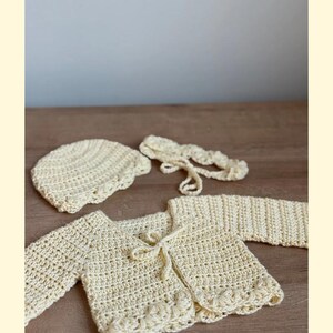 K&ouml;nnte beinhalten: Ein hellgelbes H&auml;kelset bestehend aus einer lang&auml;rmeligen Strickjacke mit Bindeband vorne und dekorativem Wellensaum, einer passenden runden Haube mit strukturiertem Muster sowie einem d&uuml;nnen, geflochtenen Stirnband mit Bindeb&auml;ndern.