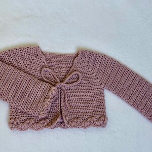 Pu&ograve; includere: Un cardigan all'uncinetto rosa antico con maniche lunghe e chiusura con fiocco. I bordi delle maniche e l'orlo inferiore presentano un design a conchiglia. Il maglione fatto a mano &egrave; esposto su una superficie bianca.