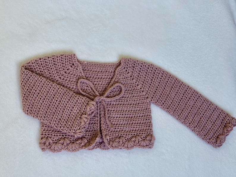 Pu&ograve; includere: Un cardigan all'uncinetto rosa antico con maniche lunghe e chiusura con fiocco. I bordi delle maniche e l'orlo inferiore presentano un design a conchiglia. Il maglione fatto a mano &egrave; esposto su una superficie bianca.