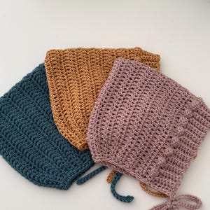 Puede incluir: Tres gorros de beb&eacute; de ganchillo en verde azulado, amarillo mostaza y malva. Cada gorro tiene una forma redondeada con un patr&oacute;n de puntada texturizada. El gorro malva tiene una fila decorativa de bolitas y lazos en la barbilla.