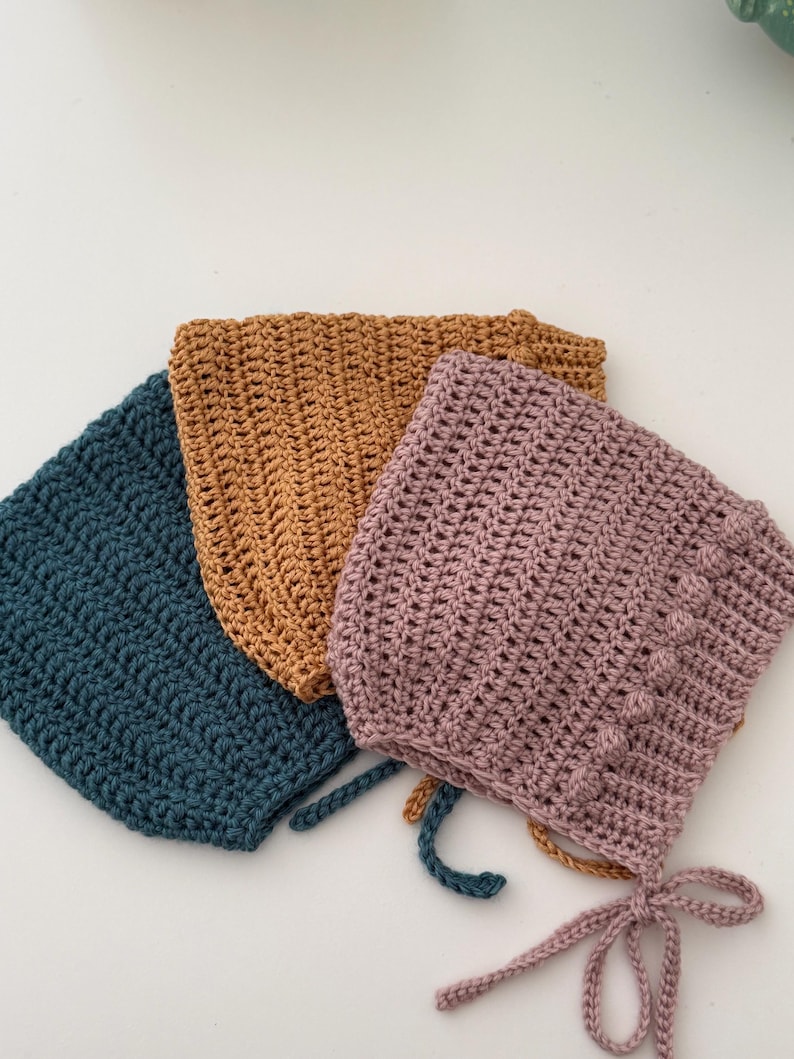 Puede incluir: Tres gorros de beb&eacute; de ganchillo en verde azulado, amarillo mostaza y malva. Cada gorro tiene una forma redondeada con un patr&oacute;n de puntada texturizada. El gorro malva tiene una fila decorativa de bolitas y lazos en la barbilla.