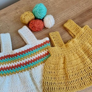 May include: Two crochet tops, one white with a colorful striped design and one yellow, are displayed with several balls of yarn in shades of yellow, orange, teal, and white. The tops have strap details and are handmade.