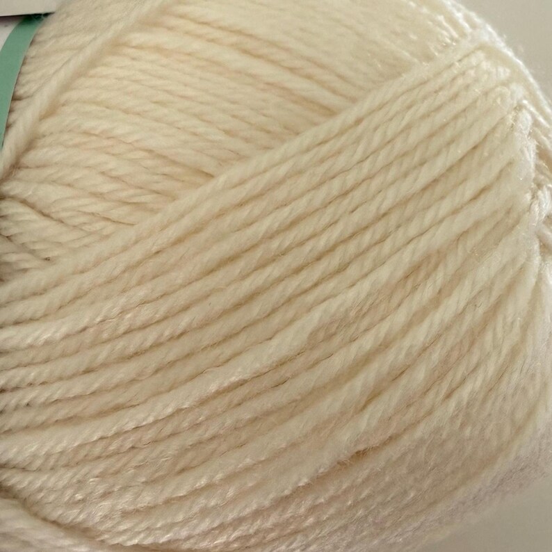 Puede incluir: Primer plano de un ovillo de lana blanquecina. La lana est&aacute; bien enrollada, con hebras visibles que crean una superficie texturizada. El color es un tono suave y cremoso, ideal para proyectos de tejido o ganchillo. La lana parece ser una fibra natural.