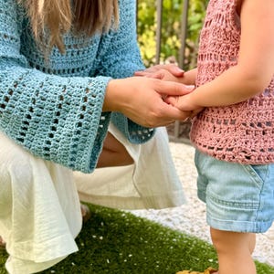 May include: A light blue long-sleeved crochet top with a wide-knit pattern, paired with white trousers. A dusty pink sleeveless crochet top with a matching wide-knit pattern, light blue denim shorts, and mustard yellow rubber sandals with buckle straps.