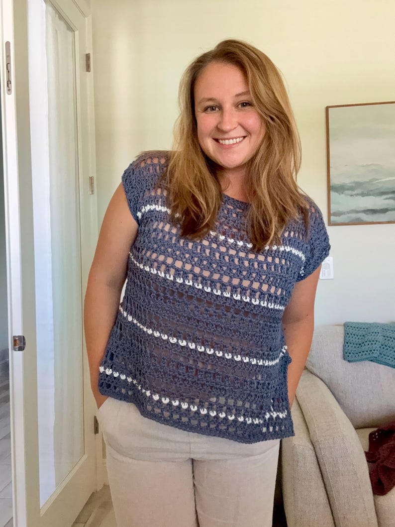 May include: A short-sleeved crochet top featuring a blue open-knit pattern with horizontal white stripes. The garment has a round neckline and a loose, boxy fit, constructed with a repeating stitch design that creates a semi-sheer, textured appearance.