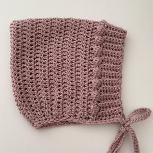Puede incluir: Un gorro de beb&eacute; tejido a ganchillo de color rosa empolvado. El gorro presenta un patr&oacute;n de puntadas texturizadas y un borde decorativo con detalles de bolitas. Tiene lazos para un ajuste seguro.