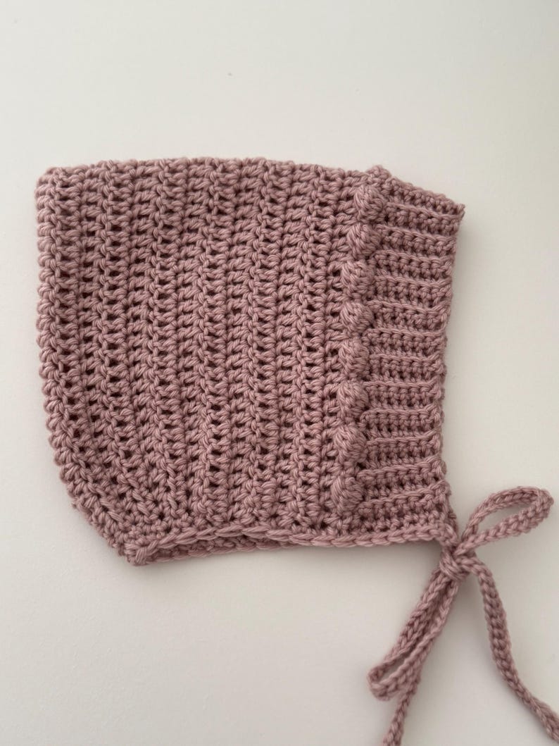 Puede incluir: Un gorro de beb&eacute; tejido a ganchillo de color rosa empolvado. El gorro presenta un patr&oacute;n de puntadas texturizadas y un borde decorativo con detalles de bolitas. Tiene lazos para un ajuste seguro.