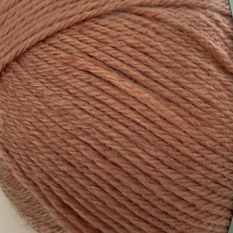 Puede incluir: Primer plano de una madeja de hilo en un tono c&aacute;lido y terroso. El hilo tiene un aspecto texturizado, con hebras visibles y una superficie ligeramente irregular. El color es un rosa apagado y polvoriento, adecuado para varios proyectos de tejido o ganchillo.
