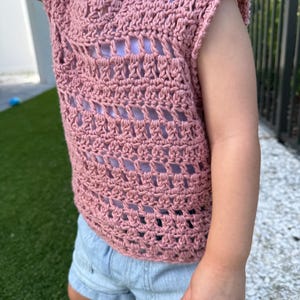 May include: A dusty rose crochet top featuring a sleeveless design with an openwork pattern and a round neckline, worn over a light purple base layer, paired with light blue denim shorts that have a cuffed hem and visible front pockets.