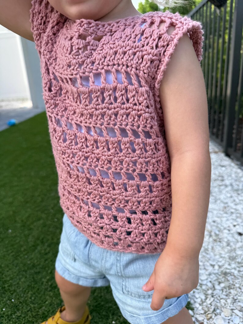 May include: A dusty rose crochet top featuring a sleeveless design with an openwork pattern and a round neckline, worn over a light purple base layer, paired with light blue denim shorts that have a cuffed hem and visible front pockets.