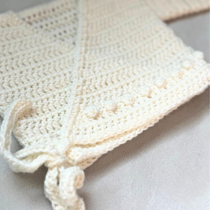 Puede incluir: Un gorro de ganchillo en color blanco roto que presenta un patr&oacute;n de puntos texturizados con una hilera de peque&ntilde;as protuberancias en relieve a lo largo del borde frontal. Incluye un cierre de lazo de ganchillo a juego en la barbilla.