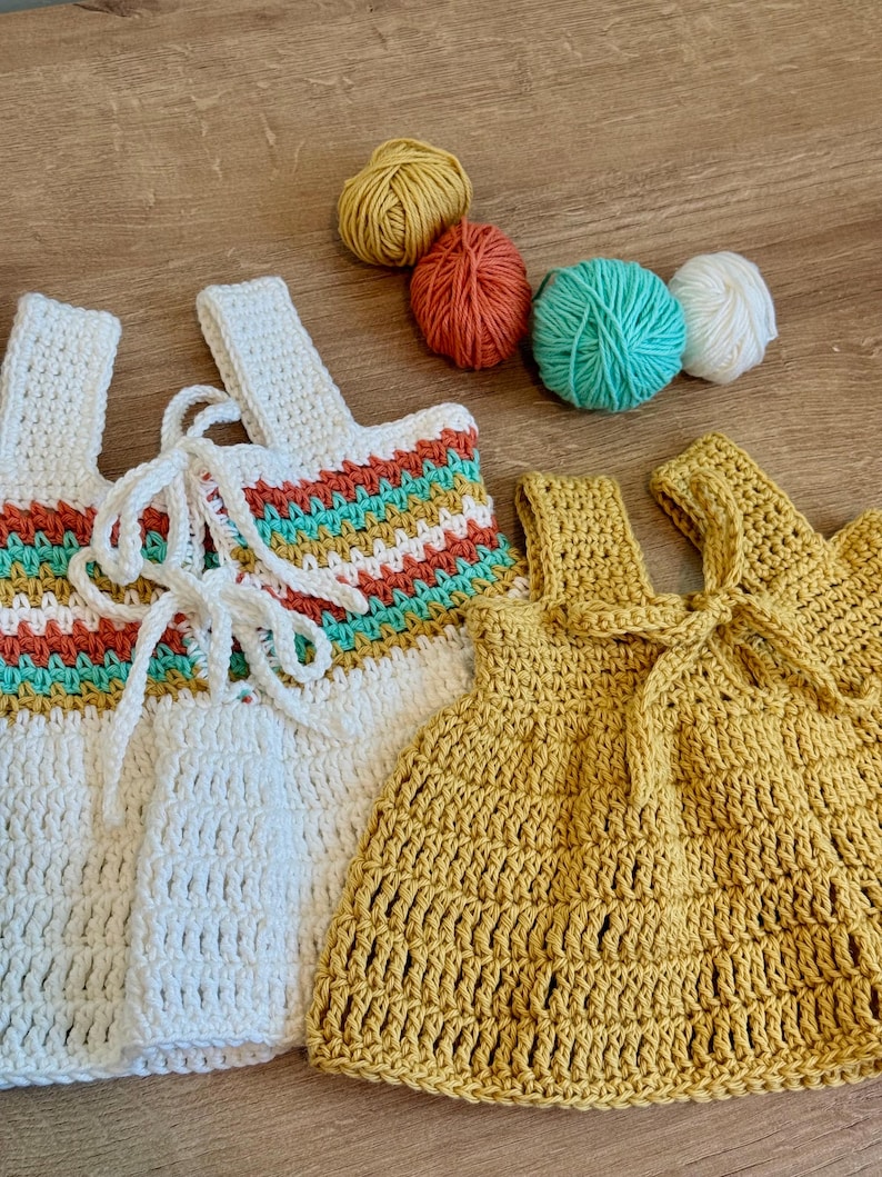 May include: Two handmade crochet tops, one white with rainbow stripes and the other yellow, are displayed. The white top has a laced front, while the yellow one has a bow. Four balls of yarn in yellow, orange, teal, and white are also visible.