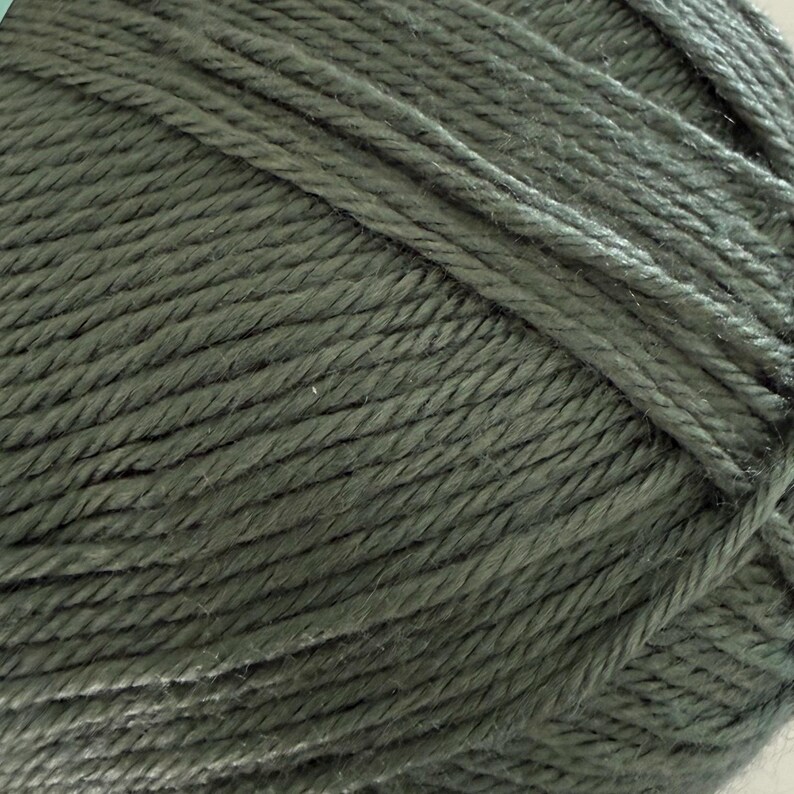 Puede incluir: Primer plano de una madeja de hilo verde oliva. El hilo est&aacute; bien enrollado, mostrando la textura de los hilos individuales. El color es un tono apagado y terroso, adecuado para varios proyectos de tejido o ganchillo.