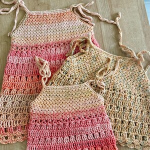 May include: Three sleeveless crochet tops featuring tie-up shoulder straps. Two tops display a gradient of peach, pink, and coral tones, while the third top features a speckled beige and brown yarn pattern with an open-work stitch design.