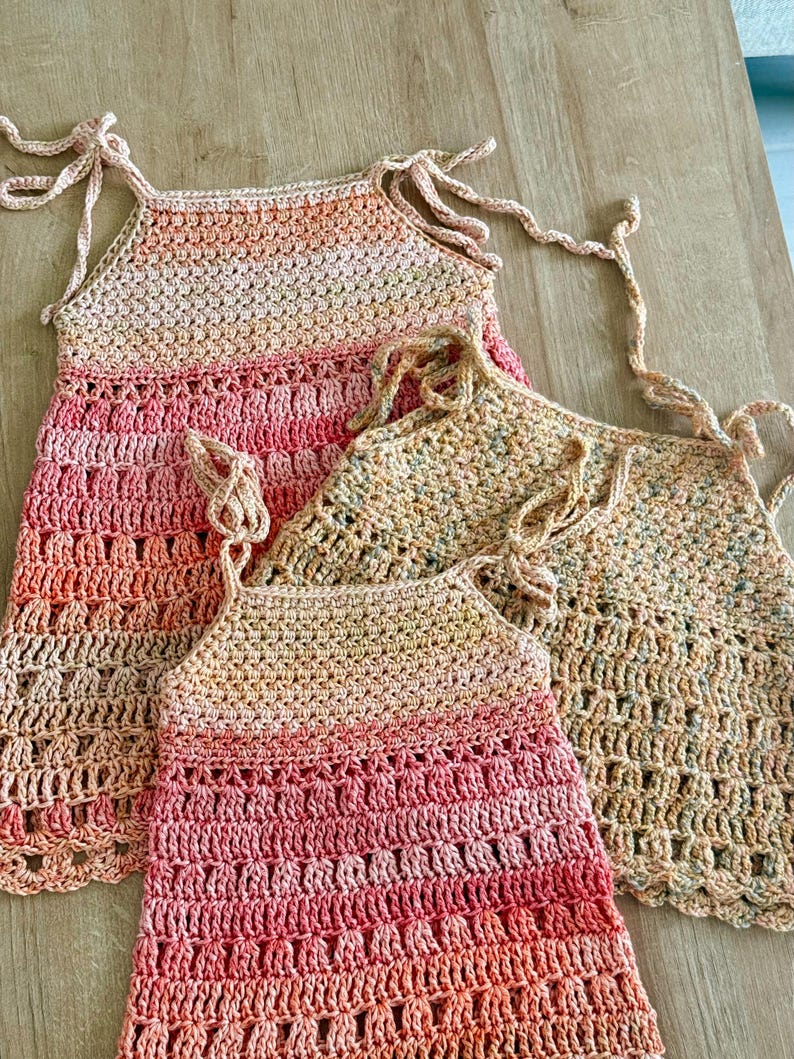 May include: Three sleeveless crochet tops featuring tie-up shoulder straps. Two tops display a gradient of peach, pink, and coral tones, while the third top features a speckled beige and brown yarn pattern with an open-work stitch design.