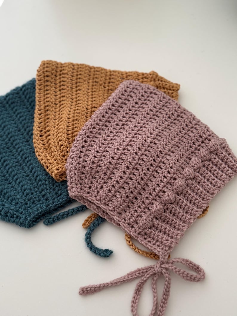Puede incluir: Tres gorros de ganchillo hechos a mano en verde azulado, marr&oacute;n y malva. Cada gorro presenta un patr&oacute;n de puntada texturizada y lazos en la barbilla. Los gorros est&aacute;n hechos de hilo suave y son adecuados para beb&eacute;s y ni&ntilde;os peque&ntilde;os.