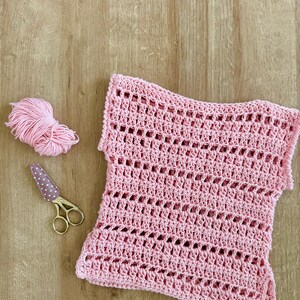 May include: A light pink crochet top featuring a repeating openwork stitch pattern, accompanied by a small ball of matching pink yarn and a pair of gold-toned embroidery scissors with a brown and white polka-dot fabric cover on the blades.